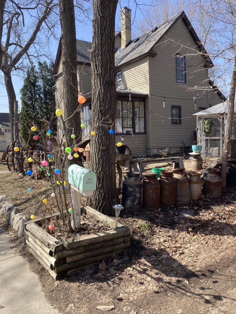 house in the background. There is a small tree in the foreground with Easter eggs on the ends of the branches. Behind the tree a statue composed of old wheels is slightly visible.