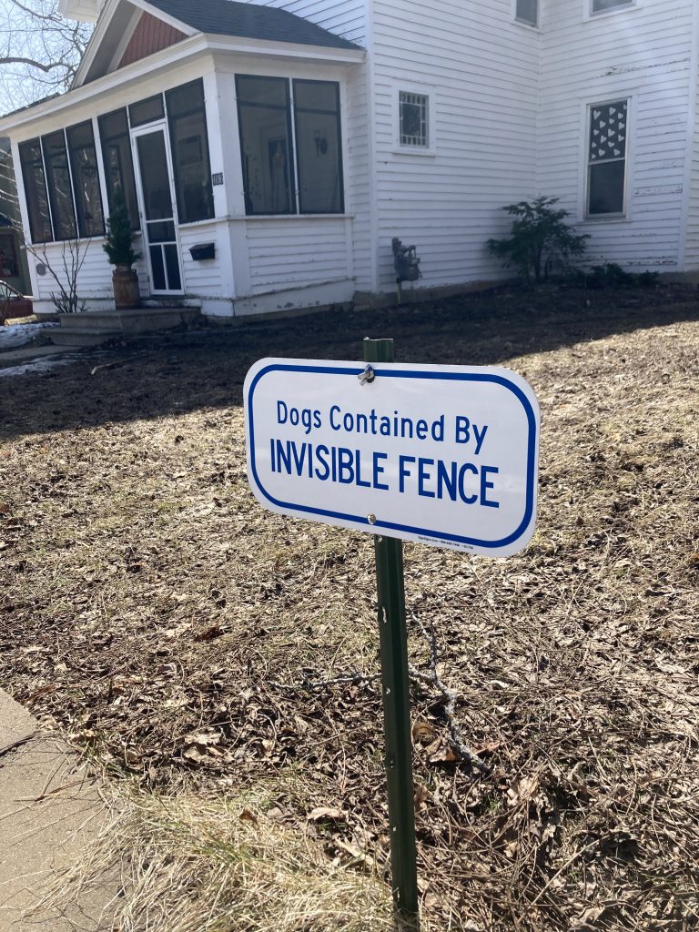 A sign fills the foreground reading "Dogs Contained By Invisible Fence". There is a white house in the background.