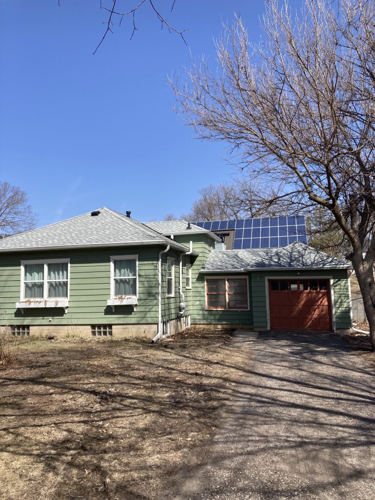 There is a one story green house in front, with a solar-paneled roof visible behind.