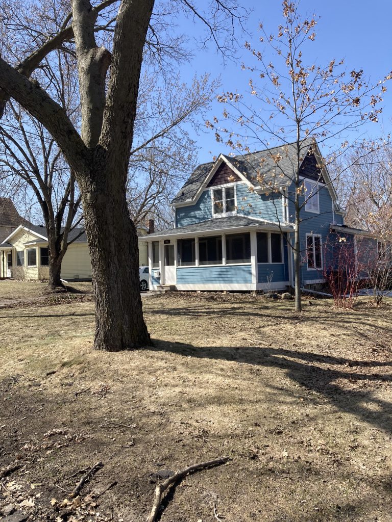 A two story blue house with white trim and a screen porch sits in a yard with some trees and brown grass.