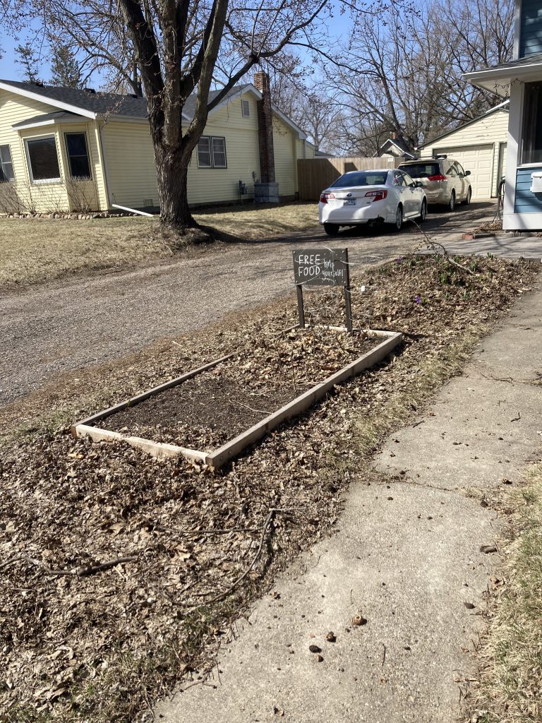 A small garden plot with the free food sign is in front of a house and driveway.