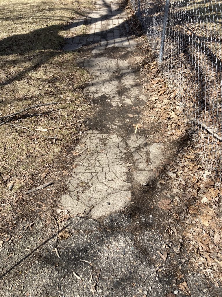 A sidewalk is broken in spidery lines and disappears into dirt in places.