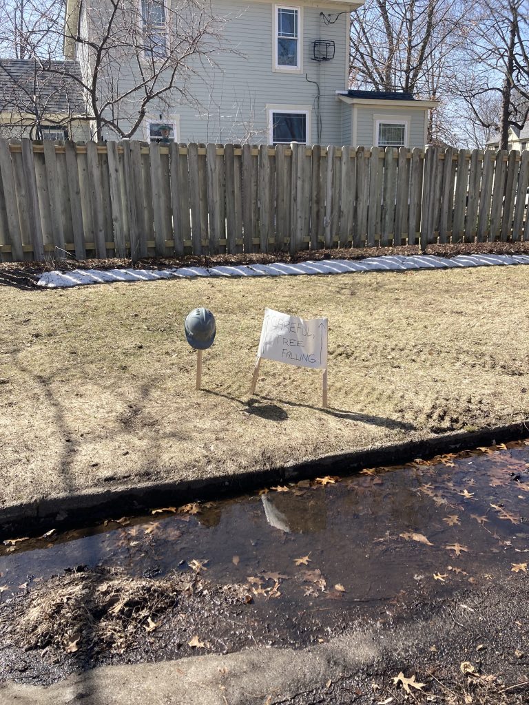 A stake in the ground with a hard hat and the sign warning about the tree. This is in front of a house that is fenced in.