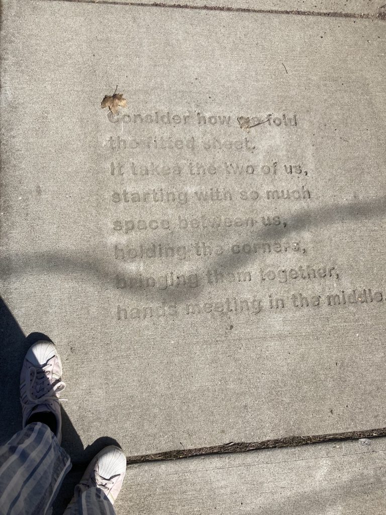 sidewalk poem reading "Consider how we fold the fitted sheet. It takes the two of us, starting with so much space between us, holding the corners, bringing them together, hands meeting in the middle.