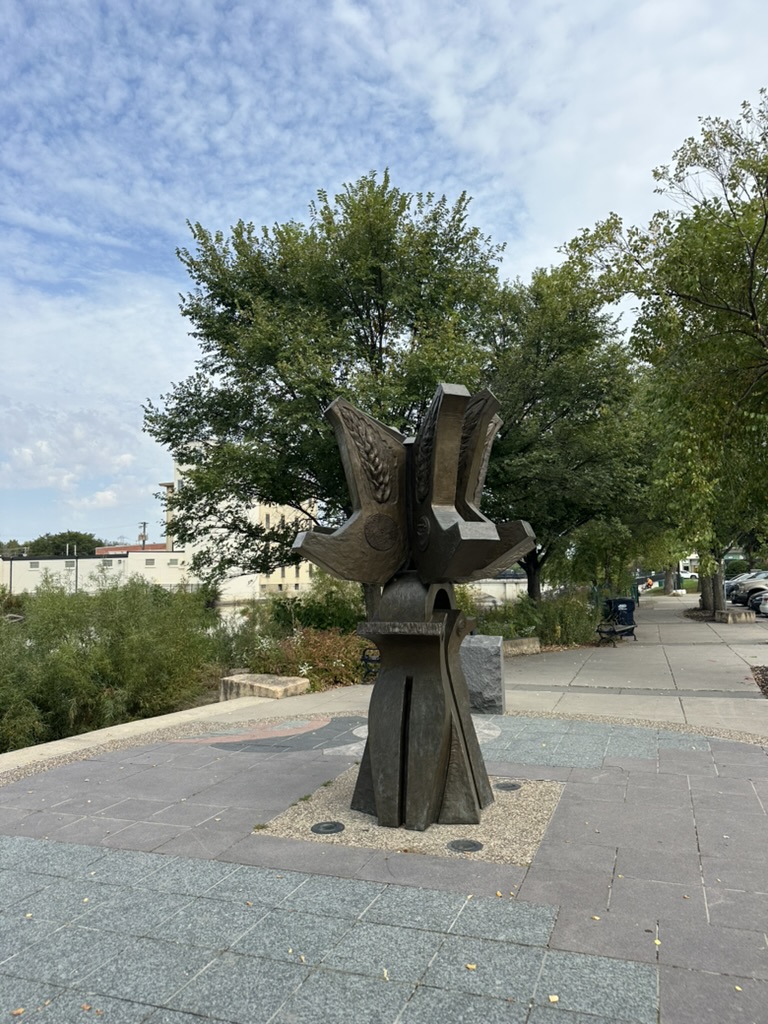 Image of bronze sculpture in front of trees and blue sky.
